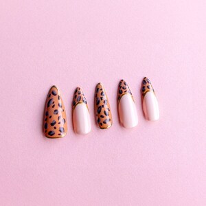 Puede incluir: Cinco uñas postizas con un diseño de estampado de leopardo. Las uñas son de color marrón claro con manchas negras y un acento dorado. Las puntas de las uñas están pintadas de blanco.