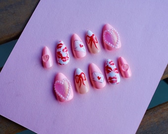 May include: Set of ten pink and white false nails with different designs. Some nails have a heart, a bow, a letter, or a red lip print.