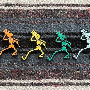 May include: A collection of six colorful skeleton figures, each wearing a top hat and holding a cane, in shades of pink, orange, yellow, green, white, and purple. The figures are arranged on a striped, textured surface.