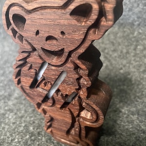 May include: A dark brown wooden cutout shaped like a dancing bear with a smiling face and rounded ears. The figure features a layered design with cut-out sections that create depth and texture, showcasing the natural wood grain throughout the piece.