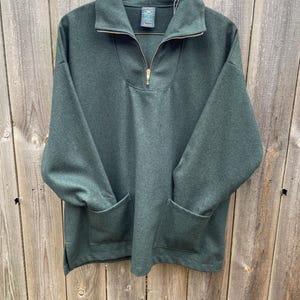 May include: A dark green pullover sweatshirt with a gold zipper and two front pockets. The sweatshirt has a high collar and is hanging on a metal hanger against a wooden fence.