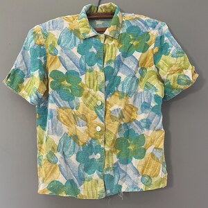 May include: Short-sleeved button-up shirt with a floral pattern in shades of blue, yellow, and green. The shirt has a classic collar and white buttons. The fabric appears to be lightweight, suitable for warmer weather.
