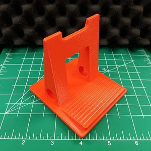 Puede incluir: Un soporte naranja brillante impreso en 3D. Presenta una base rectangular con una superficie ranurada y una estructura vertical con una abertura rectangular. El soporte está sobre una esterilla de corte verde con medidas en centímetros.