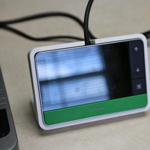 Könnte beinhalten: Ein rechteckiger digitaler Kontrollbildschirm mit weißem Rahmen und grünem Sockel, der den Text xTool trägt. Das Gerät verfügt über ein schwarzes Display, ein schwarzes Kabel an der Oberseite und ein vertikales Bedienfeld mit Pfeilen rechts.