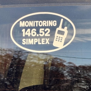 Könnte beinhalten: Weißer ovaler Aufkleber mit der Aufschrift "MONITORING 146.52 SIMPLEX" und einer Walkie-Talkie-Silhouette. Der Aufkleber befindet sich auf einer Autoscheibe, mit Reflexionen.