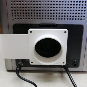 Peut inclure: Un appareil électronique argenté doté d'un panneau supérieur perforé et d'une section centrale noire. Un adaptateur d'échappement en plastique blanc de forme carrée avec une ouverture circulaire est fixé à l'arrière, s'étendant vers la gauche.