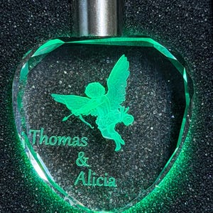 May include: A heart-shaped clear glass pendant featuring an internal laser-etched green glowing image of a winged cherub holding a bow and arrow. The text "Thomas & Alicia" is engraved below the cherub. The pendant has a silver-toned metal cap.
