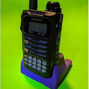 Könnte beinhalten: Ein schwarzes digitales Yaesu FT-70D Handfunkgerät mit Antenne, Monochrom-Display und Ziffernblock, das in einer violetten Kunststoff-Ladestation steht. Das Gerät verfügt über beschriftete Tasten sowie Anschlüsse für Mikrofon, Daten und externe Stromversorgung.