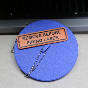 Peut inclure: Un couvercle rond en plastique bleu avec une petite poignée au centre. Attachée au couvercle par une chaînette à billes en métal argenté, une étiquette rectangulaire orange avec une bordure noire porte l'inscription REMOVE BEFORE FIRING LASER.