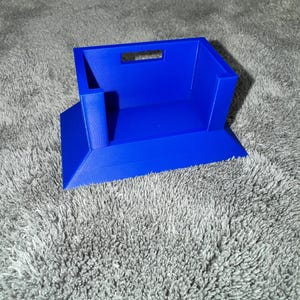 Puede incluir: Un organizador azul brillante, impreso en 3D, con forma rectangular y un recorte para el asa. El organizador tiene una base inclinada y un pequeño soporte vertical en un lado. El objeto está sobre una superficie gris texturizada.