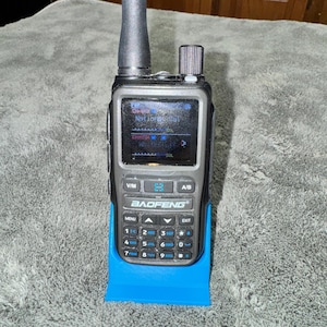 Puede incluir: Una radio Baofeng negra y gris con una pantalla que muestra texto, colocada en un soporte azul brillante. La radio tiene una antena negra y una serie de botones y un teclado. La pantalla muestra el texto "Nart Jorgical".