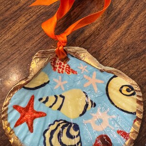 May include: A seashell ornament with a blue background and a gold rim. The shell is decorated with painted seashells, starfish, and coral in shades of red, yellow, and white.