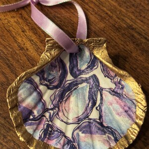 May include: A decorative seashell ornament with a gold-painted edge and a purple ribbon for hanging. The shell is filled with a watercolor design in shades of purple, blue, and pink, creating a coastal theme.