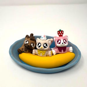 May include: A whimsical dessert-themed figurine set featuring three cat figures in a blue bowl. The cats are brown, tan, and pink, each with unique details like a chocolate bar, a party hat, and a strawberry. They sit on a yellow banana.