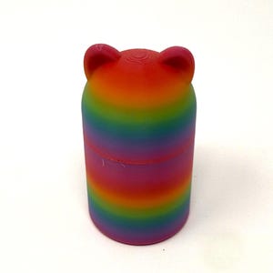 May include: A cylindrical container with a rainbow gradient design, featuring a bear-shaped top with two small ears. The colors transition from red to purple. The container is made of a solid material.