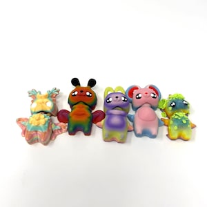 May include: A collection of six colorful, whimsical figurines. Each figure has a unique design, with variations in color and shape. The figurines are arranged in a row, showcasing their diverse features and vibrant hues. The overall theme is playful and imaginative.