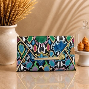 May include: A rectangular envelope clutch bag featuring a vibrant, multi-colored snakeskin pattern in shades of green, blue, yellow, pink, and black. The bag has a pointed flap closure accented with a thin, horizontal gold-toned metal bar.