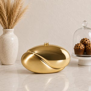 May include: A polished gold-toned, oval-shaped container with a smooth, reflective surface and a small, circular knob on top. The object features a curved, asymmetrical design that separates into two distinct sections, resting on a flat surface.
