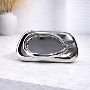 Puede incluir: Una bandeja decorativa plateada brillante con un diseño de asa curvada único. La bandeja tiene una superficie lisa y reflectante y un interior gris oscuro. Se coloca sobre una superficie de mármol blanco.