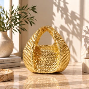 May include: A metallic gold handbag featuring a woven texture and a rounded, integrated handle design. The bag has a structured, curved silhouette with a consistent braided pattern throughout the exterior surface, reflecting light across its surface.