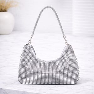 Könnte beinhalten: Eine silberne Handtasche mit geschwungener Form und Kettenriemen. Die Tasche ist mit kleinen, glitzernden Strasssteinen bedeckt, was ihr ein glamouröses Aussehen verleiht. Der Griff ist ebenfalls aus Strass gefertigt. Die Tasche steht auf einer weißen Oberfläche.