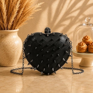 May include: A black heart-shaped clutch bag featuring a textured surface adorned with numerous dark metal spikes. The bag includes a skull-shaped clasp at the top center and a thin black chain shoulder strap attached to both sides of the frame.