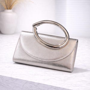 Puede incluir: Un clutch plateado con un asa grande, circular y plateada. El clutch tiene una solapa curva y un acabado metálico liso. El asa está sujeta en la parte superior. El fondo es blanco suave.