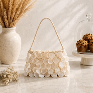 Peut inclure: Un sac à main doté d'un corps structuré recouvert de disques de coquillages blancs circulaires superposés. Le sac possède une anse tressée unique fixée sur le dessus, créant un aspect texturé et superposé dans des tons blanc cassé.