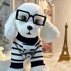 May include: White plush toy dog wearing black framed glasses, a black and white striped shirt, and a silver chain with a miniature camera. The dog is standing on a white fluffy surface with a gold Eiffel Tower in the background.