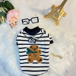 May include: A black and white striped dog shirt with a brown teddy bear applique and a small camera charm on a silver chain.