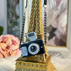 May include: A miniature black camera with a silver lens and a flash, hanging from a black chain on a gold Eiffel Tower replica.