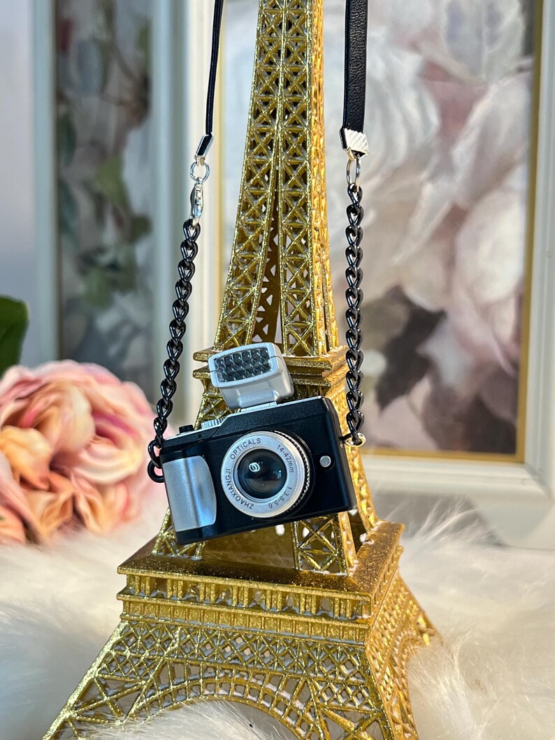 May include: A miniature black camera with a silver lens and a flash, hanging from a black chain on a gold Eiffel Tower replica.