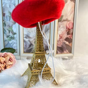 Pu&ograve; includere: Un berretto di feltro rosso appeso a una replica dorata della Torre Eiffel. Il berretto &egrave; sospeso da uno spago bianco.