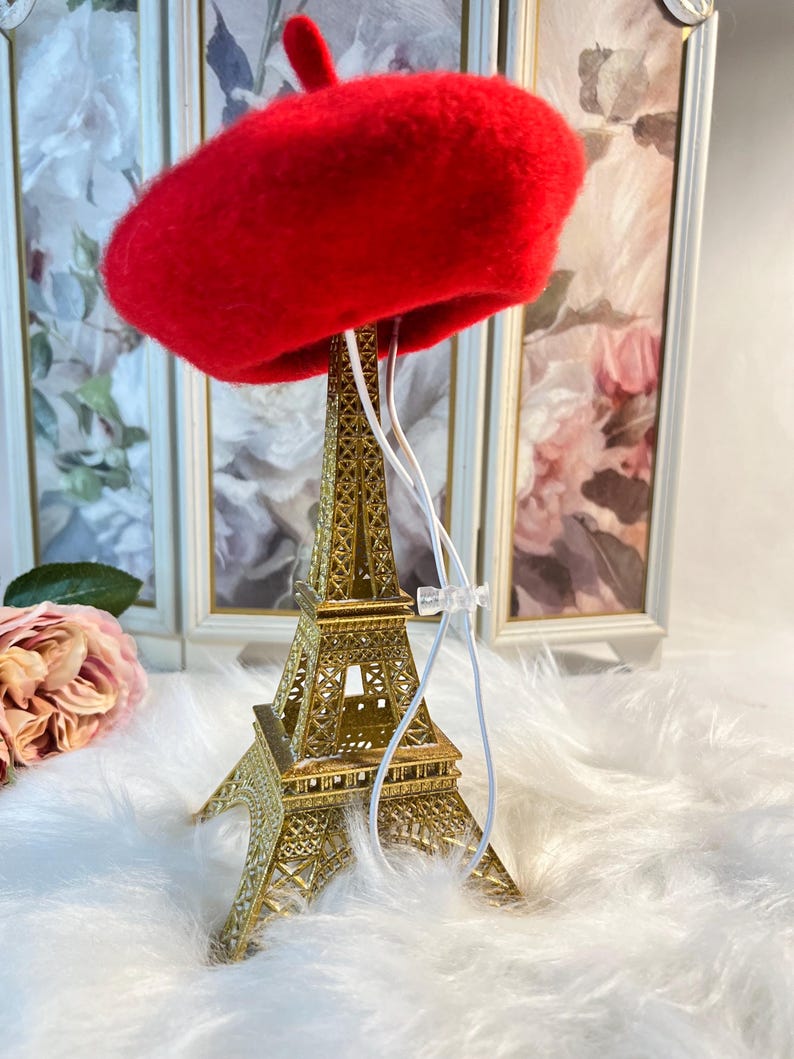 Pu&ograve; includere: Un berretto di feltro rosso appeso a una replica dorata della Torre Eiffel. Il berretto &egrave; sospeso da uno spago bianco.