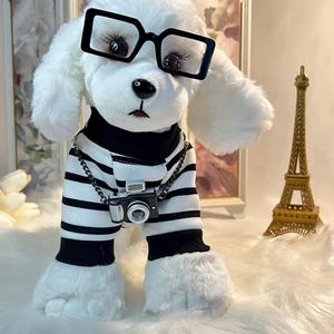 May include: White plush toy dog wearing black and white striped pyjamas, black framed glasses, and a silver chain with a miniature camera. The dog is standing on a white fluffy surface with a gold Eiffel Tower in the background.