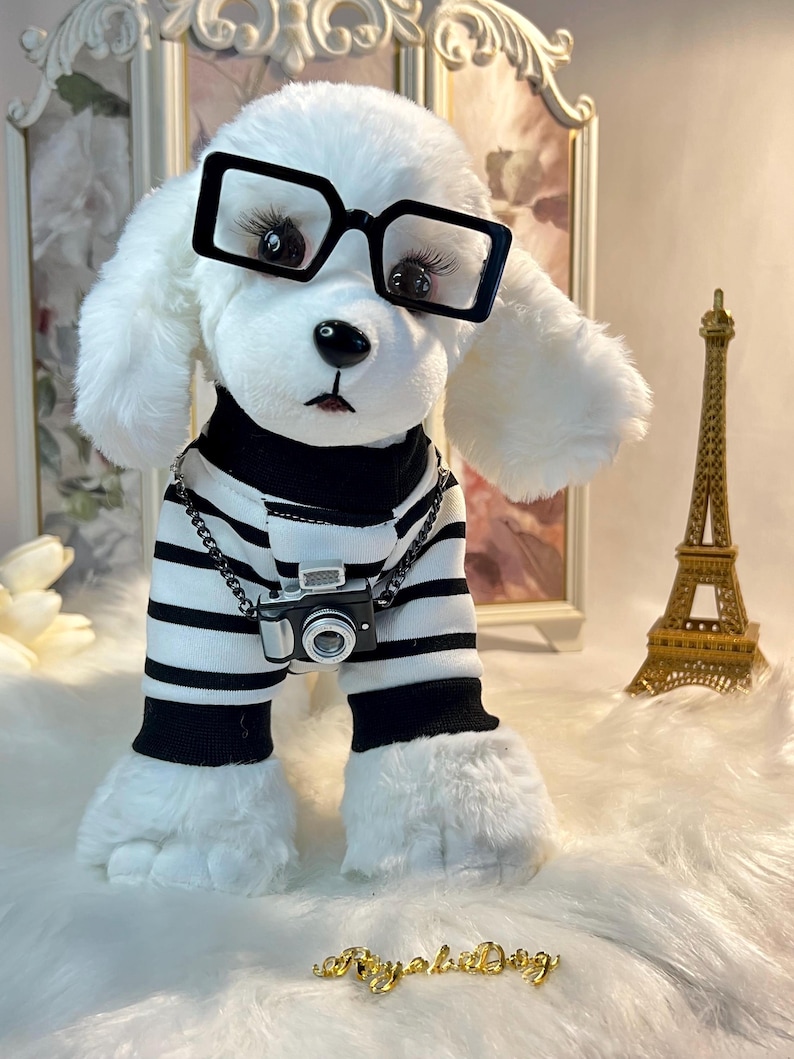 May include: White plush toy dog wearing black and white striped pyjamas, black framed glasses, and a silver chain with a miniature camera. The dog is standing on a white fluffy surface with a gold Eiffel Tower in the background.