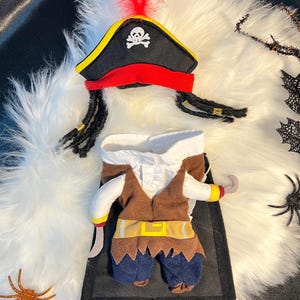 May include: A brown and blue pirate costume for a small dog, with a black hat with a skull and crossbones, a red feather, and a gold trim. The costume includes a brown vest, a white shirt, blue pants, and a hook.