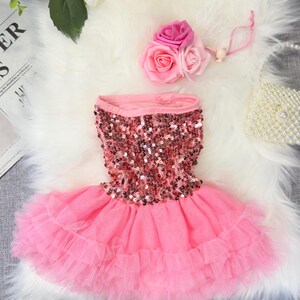 May include: Pink dog dress with a sequined bodice and layered tulle skirt. A matching pink flower hair accessory is included. The dress is designed for small dogs and is displayed on a white faux fur surface.