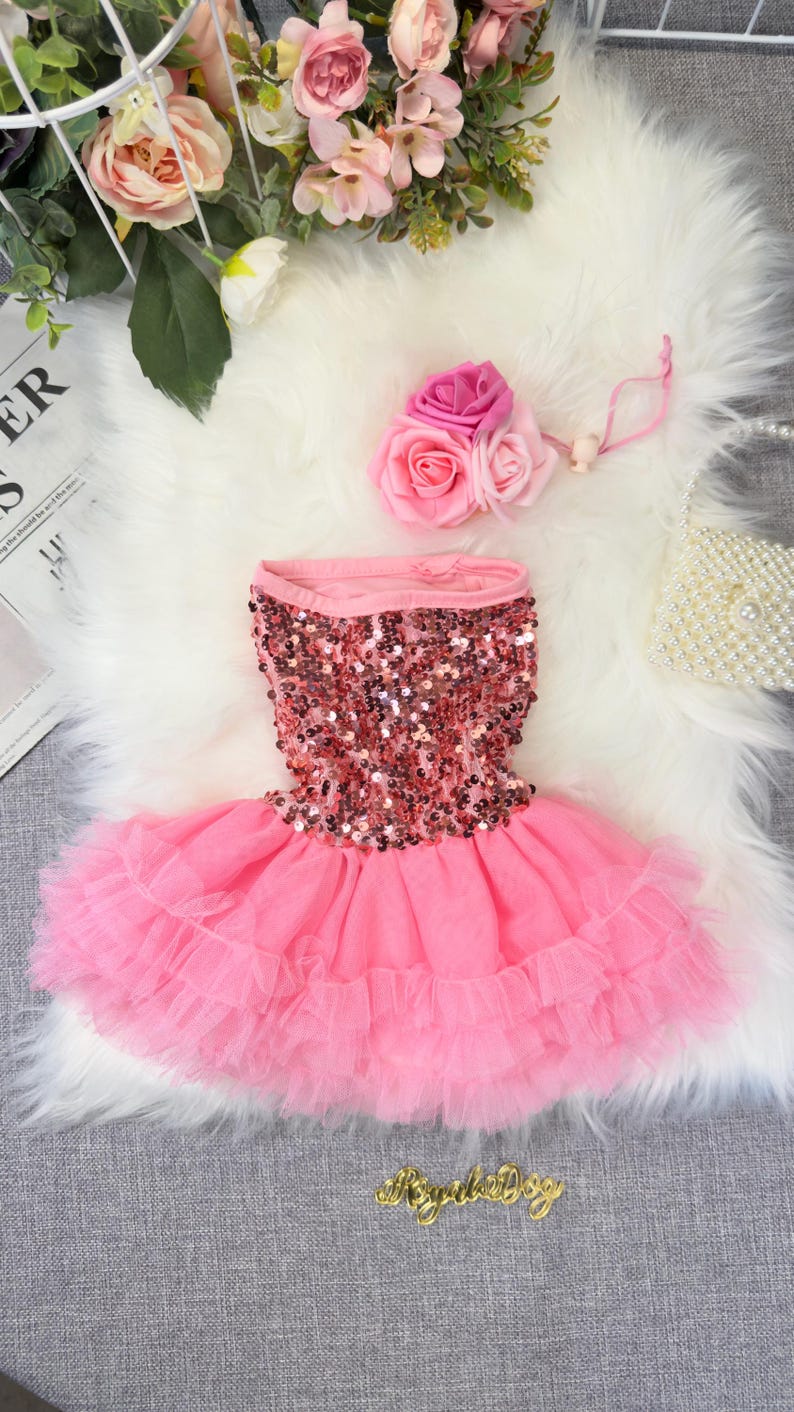 May include: Pink dog dress with a sequined bodice and layered tulle skirt. A matching pink flower hair accessory is included. The dress is designed for small dogs and is displayed on a white faux fur surface.