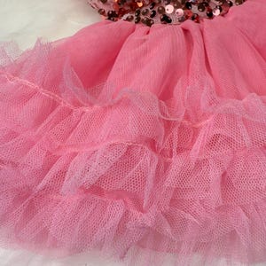 May include: A pink dress with a bodice covered in bronze sequins and a layered tulle skirt. The dress is designed for a child and is set against a white, fluffy background.