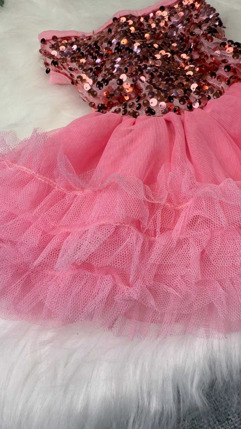 May include: A pink dress with a bodice covered in bronze sequins and a layered tulle skirt. The dress is designed for a child and is set against a white, fluffy background.