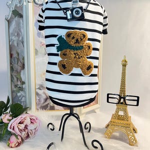 May include: A white and black striped dog shirt with a brown teddy bear applique and a black camera charm on a black chain. The shirt is on a black stand and there is a gold Eiffel Tower and black glasses in the background.