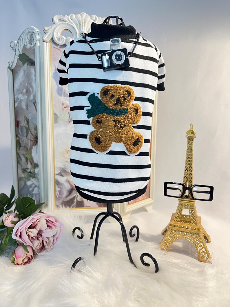 May include: A white and black striped dog shirt with a brown teddy bear applique and a black camera charm on a black chain. The shirt is on a black stand and there is a gold Eiffel Tower and black glasses in the background.