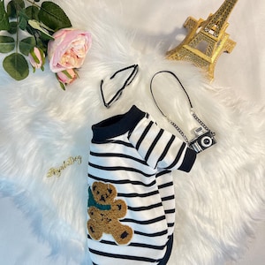 May include: A black and white striped dog shirt with a brown teddy bear applique. The shirt has a black collar and short sleeves.