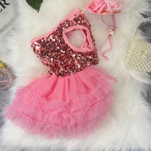May include: A pink dog dress with a sequined bodice and a layered tulle skirt. A matching pink flower headband and a small beaded purse are also present. The dress is designed for a pet and is displayed on a white fluffy surface.
