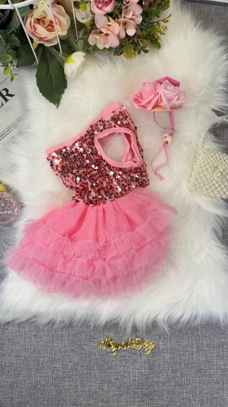 May include: A pink dog dress with a sequined bodice and a layered tulle skirt. A matching pink flower headband and a small beaded purse are also present. The dress is designed for a pet and is displayed on a white fluffy surface.