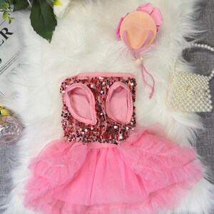 May include: A pink sequined dog dress with a layered tulle skirt. The dress has openings for the front legs and is adorned with a matching pink bow. A small pearl handbag and a pink hat are also present.