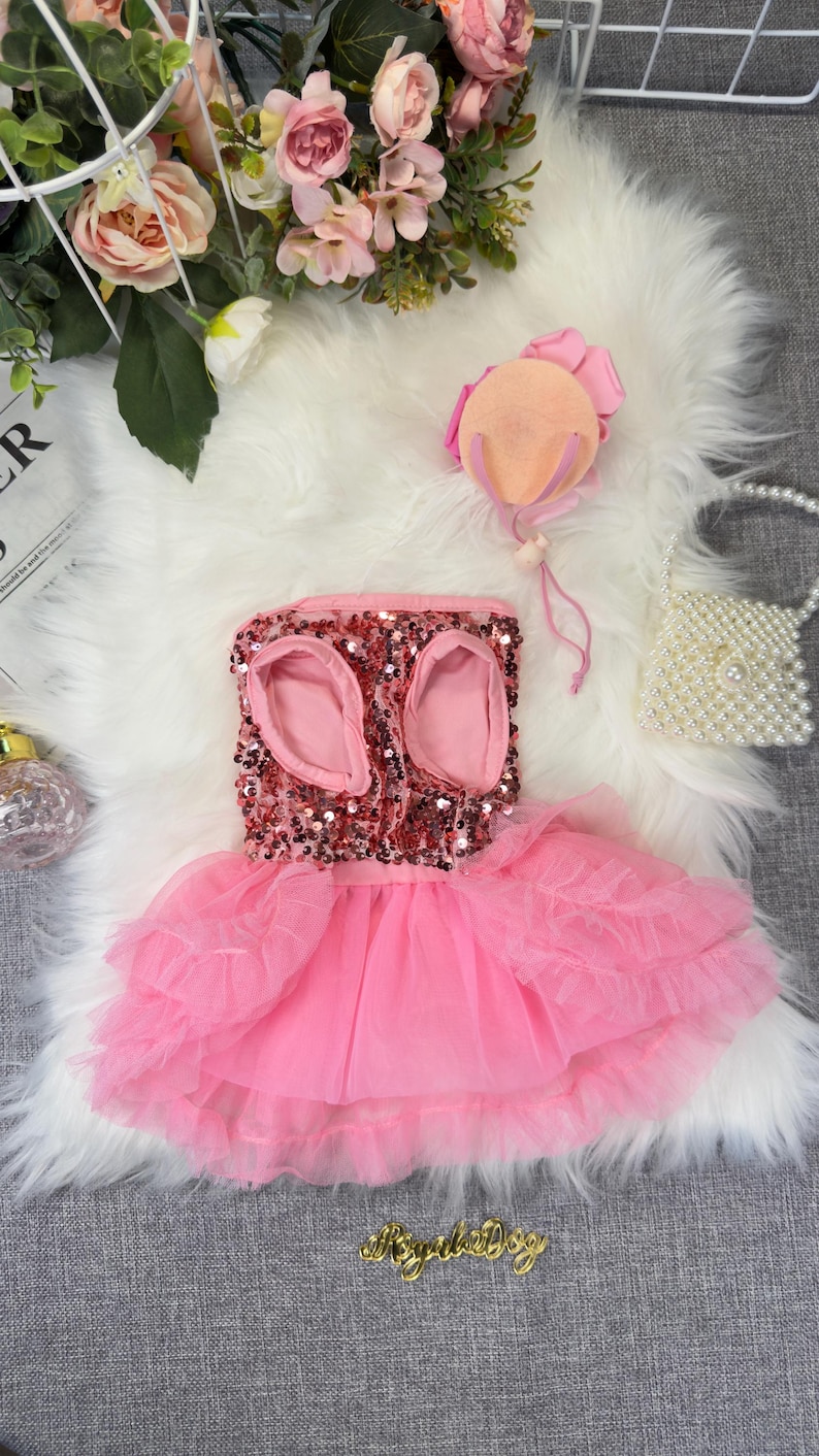 May include: A pink sequined dog dress with a layered tulle skirt. The dress has openings for the front legs and is adorned with a matching pink bow. A small pearl handbag and a pink hat are also present.