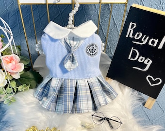 Può includere: Un vestito da cane a quadri blu e bianchi con un colletto bianco, un maglione blu e una cravatta blu. Il vestito ha una piccola toppa ricamata con il testo "Royal High School" e una corona. Il vestito è appeso a un appendiabiti di perle bianche. Il testo "Royal Dog" è scritto su una lavagna nera.