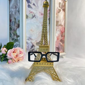 May include: Black square-shaped eyeglasses with a gold Eiffel Tower miniature in the background. The Eiffel Tower is on a white fluffy surface.